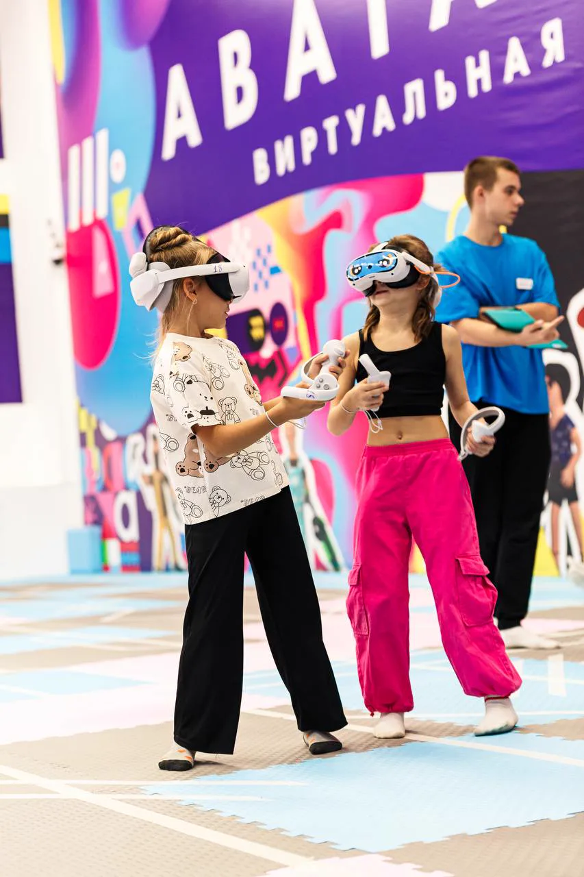 Две девочки в VR-шлемах играют на арене, рядом инструктор парка